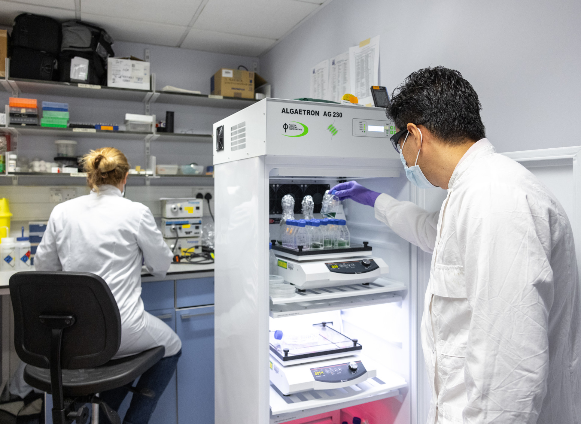 A scientist sits at a desk while another looks at flasks in a refridgerator