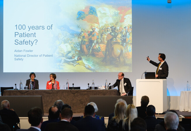 Man presents at lectern in front of slide that says "100 years of Patient Safety?"