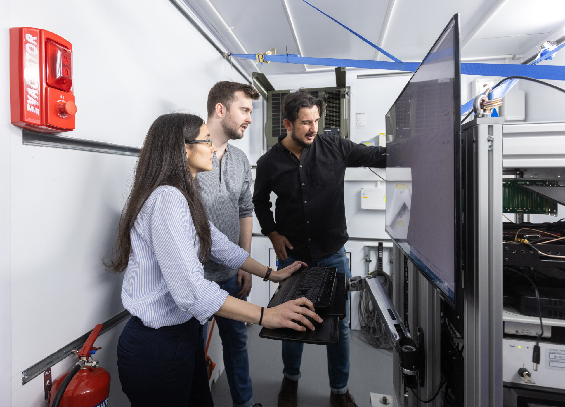 Three people looking at a screen in front of a box of wires