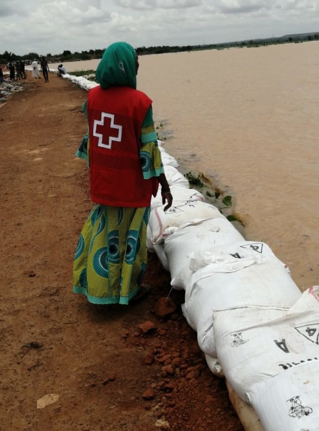 Woman wearing a vest with a red cross stands next to a river with its banks built up with sandbags