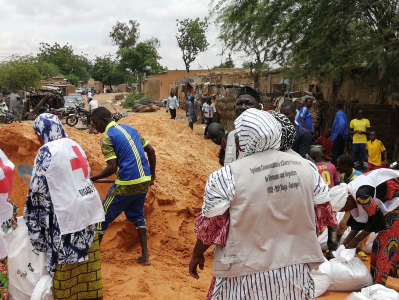 Group of people wearing red cross vests over their everyday clothes prepare sandbags, next to a big pile of sand