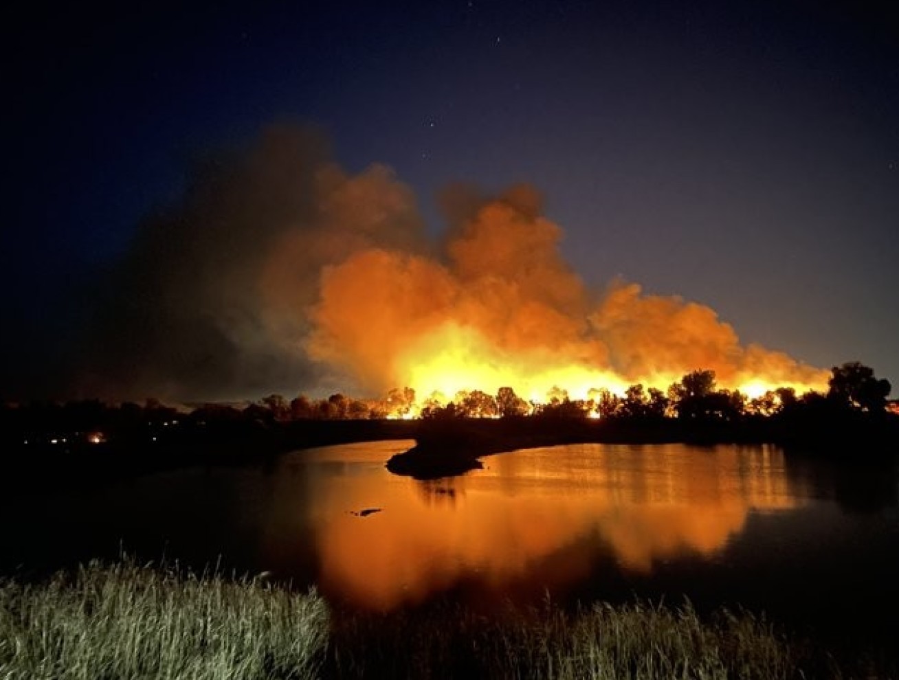 Photo: At night, a wildfire rages in the distance, with a lake in the foreground