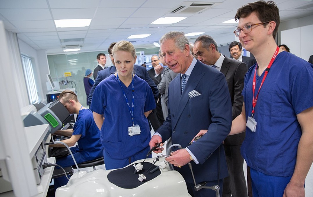 70 King Charles, with two people in blue scrubs, uses a laparoscopic simulator