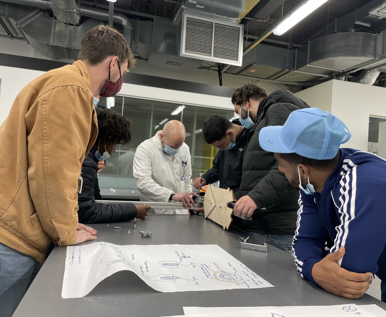 5 male teenagers sat around a table as 2 of them help build their e-scooter with a researcher in a white lab coat