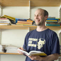 Prof Kevin Buzzard in his office holding an open book standing in front of shelves full of mathematics books and papers