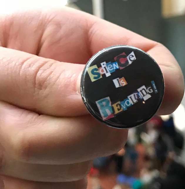 A hand holding a badge with 'Science is revolting' on it