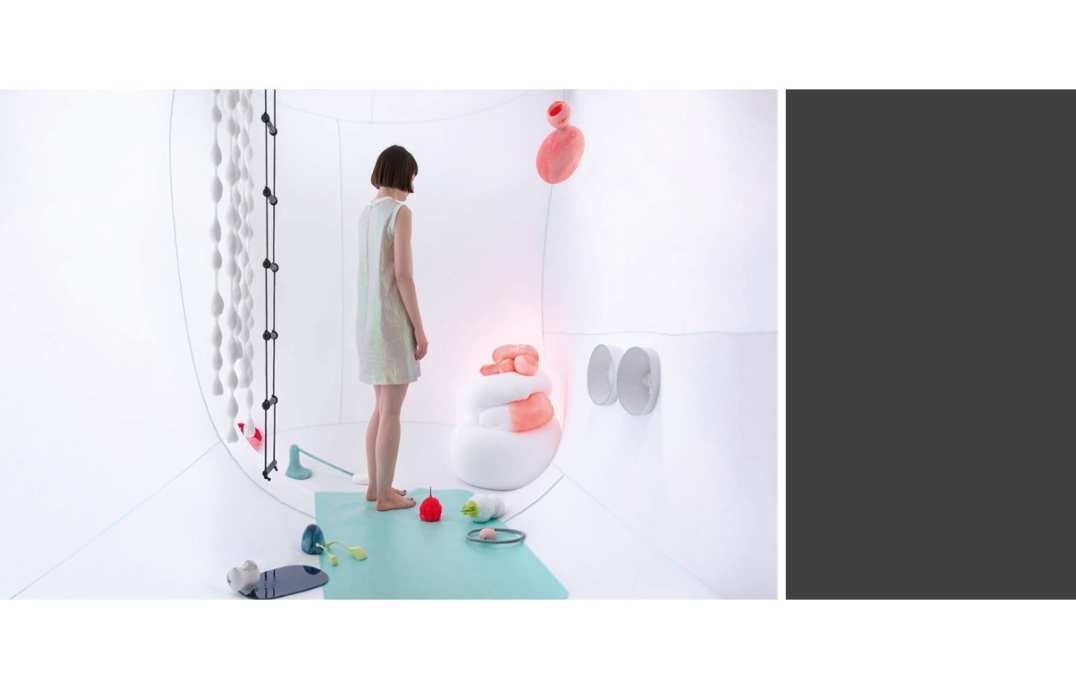 A woman stands in a white room on top of a blue mat surrounded by abstract plastic objects that look remotely like organs, to her left hangs a ladder and some large white beads,