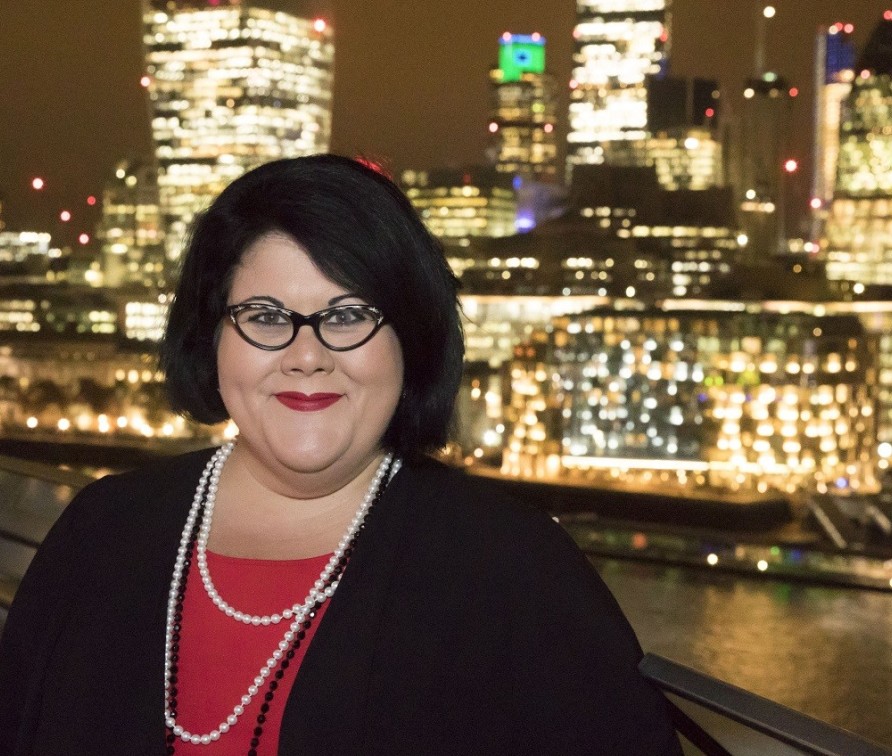 Photo of Amy Lame with black hair and a pair of black glasses, dressed in red with a black cardigan and a white necklace, against the London night sky