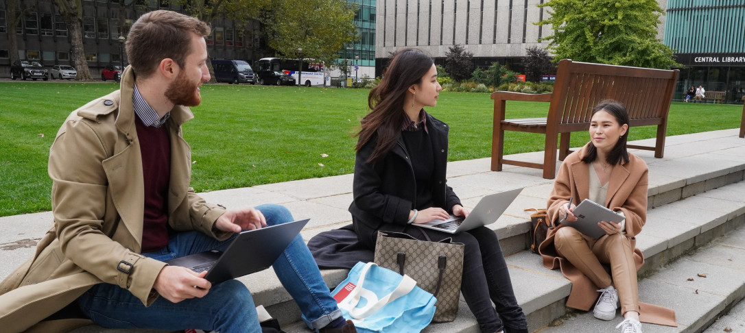 An image showing three students sitting on stone steps in front of a grass lawn working on laptops and having a conversation