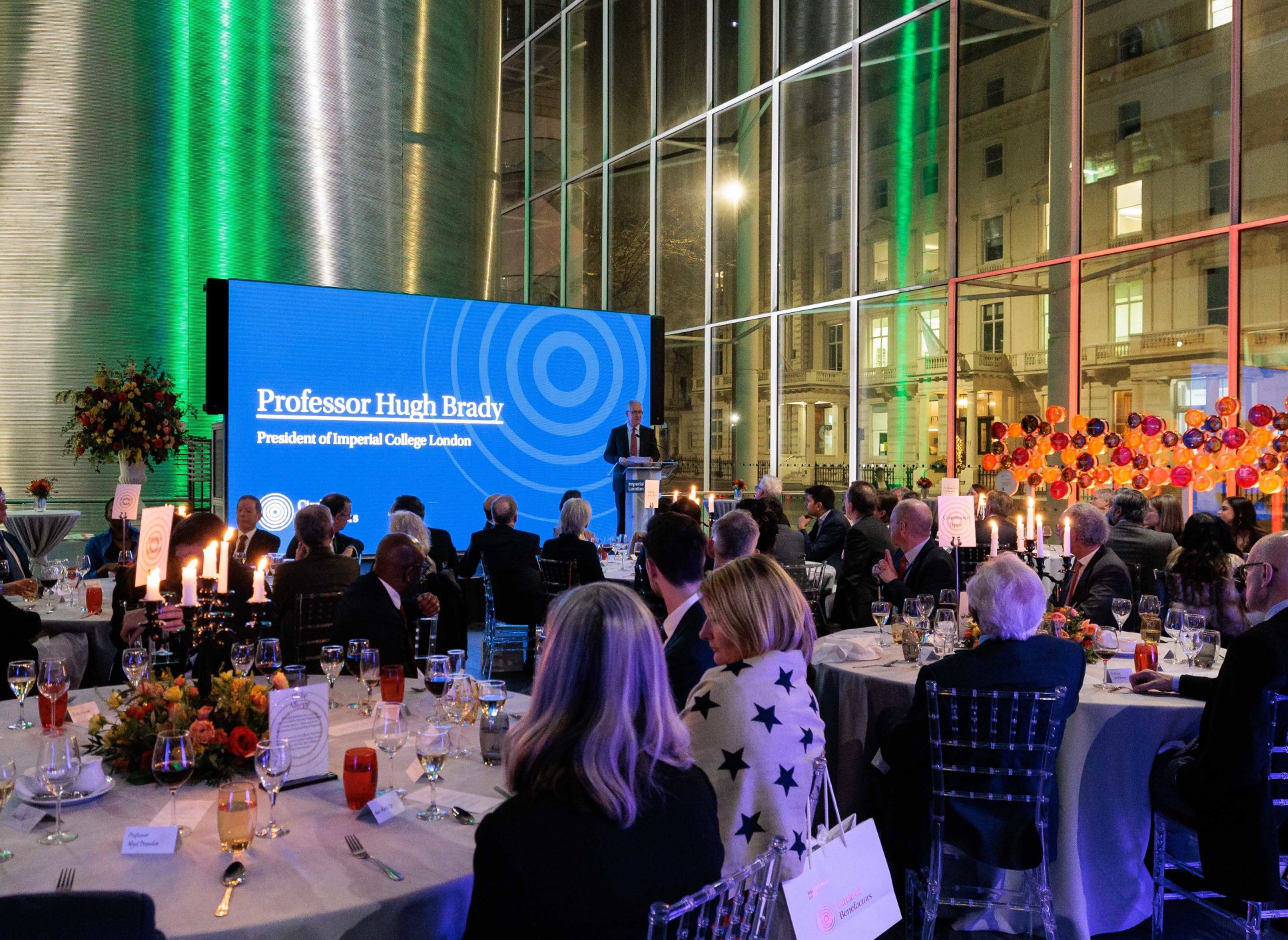 President Hugh Brady delivers a speech on a podium in front of the screen in College Main Entrance. To the right is the new donor installation and in the foreground are guests seated at tables