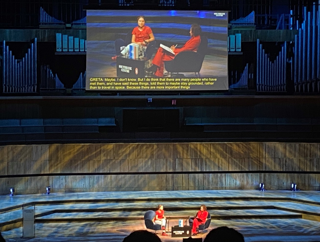 Greta Thunberg and another person sit on a stage with wood panelling, a large screen behind them relays a close-up video