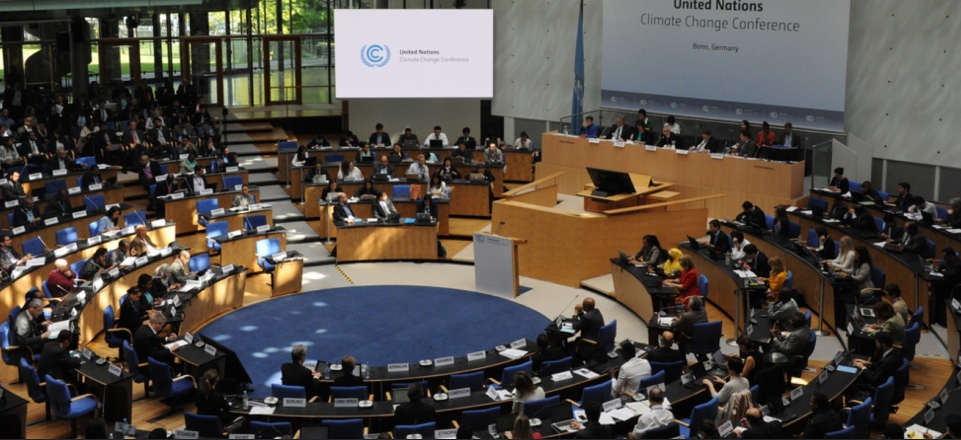 Photo shows a conference all with wooden desks, with smartly dressed people sitting. Large screen reads: United Nations Climate Change Conference, Bonn, Germany