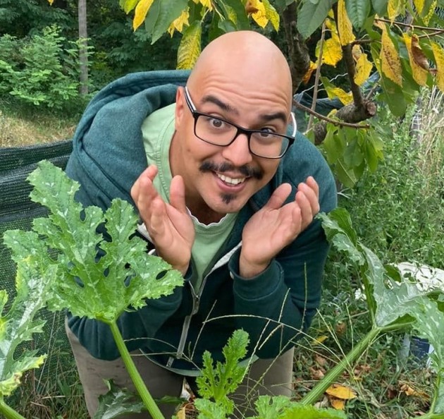 Dan, a man in his 30s with tan skin, crouches down in a garden next to a courgette plant