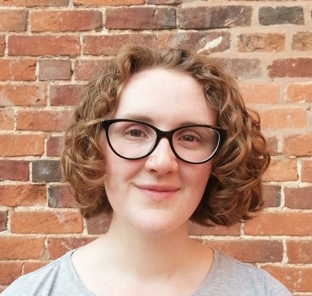 Emma, a smiling woman in her 20s with white skin and curly red hair stands in front of a brick wall