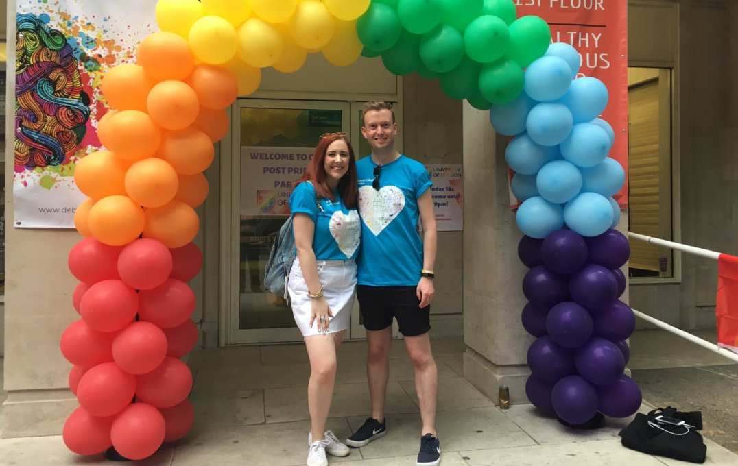 two people stand under rainbow-coloured balloon arch