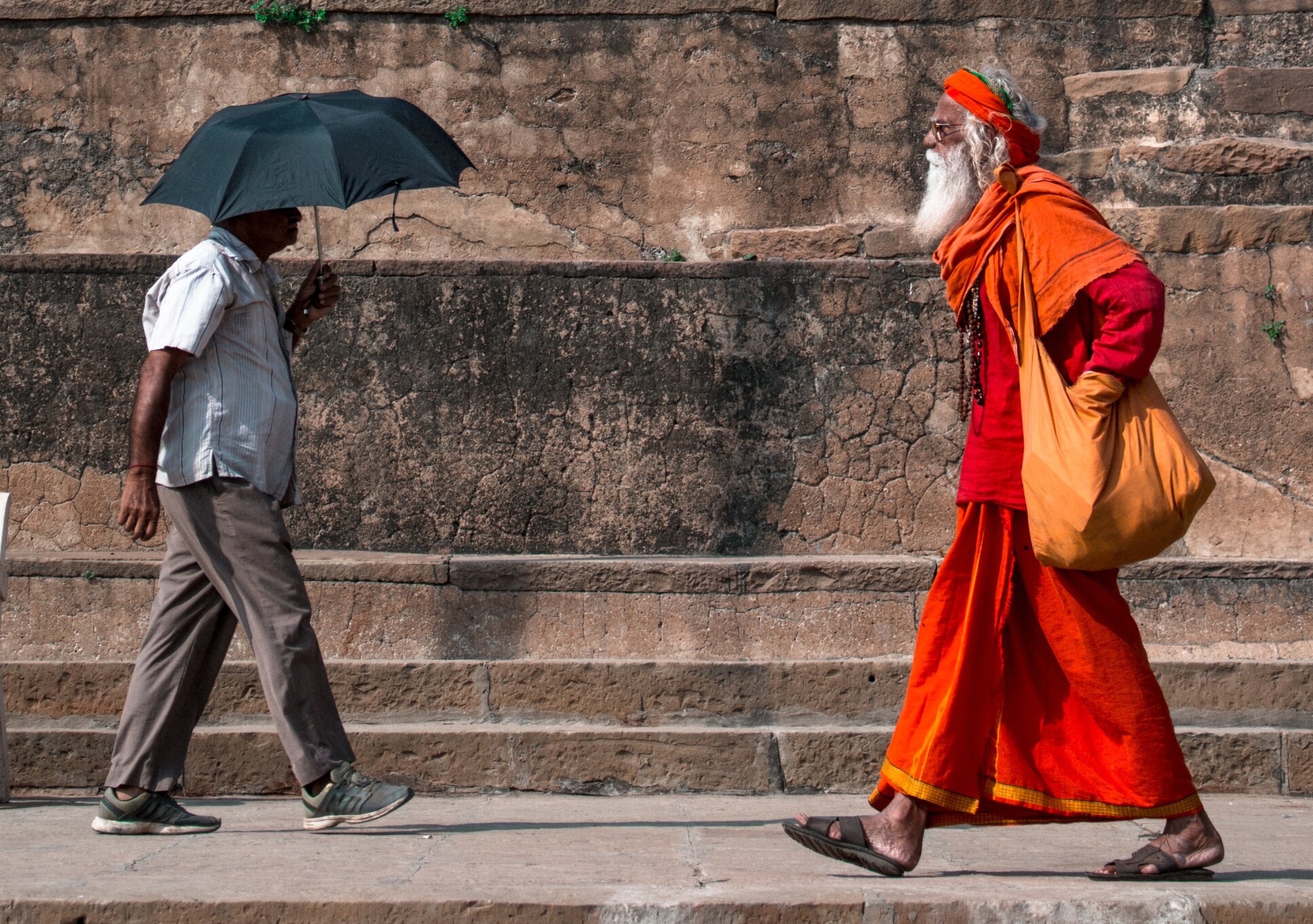 Two Indian people walk past each other on hot day, one wearing a shirt and pants with an umbrella and the other wearing orange robes.