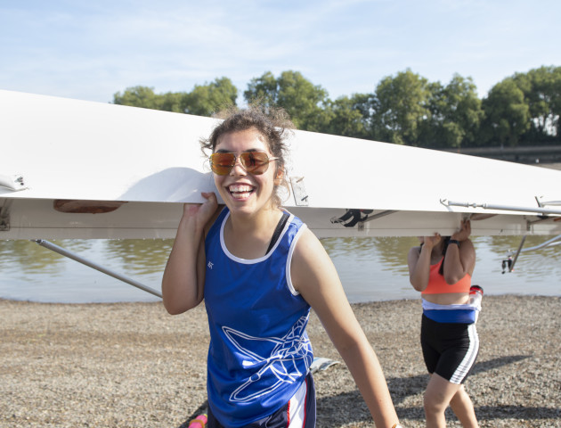 A woman rower smiles at the camera while carrying a rowing boat