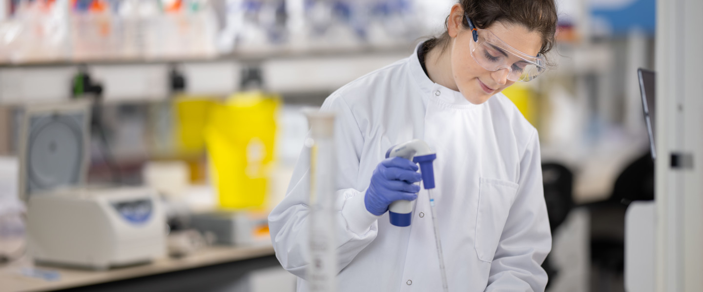 A student in a lab wearing goggles, a lab coat and surgical gloves