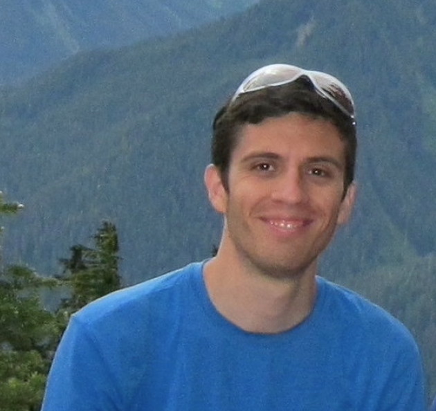 Paulo, a white man in his thirties with short hair sits on a log atop a mountain