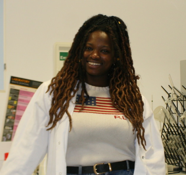 Lara, a smiling Black woman in her 20s with long braided hair. She stands in lab and wears a white coat