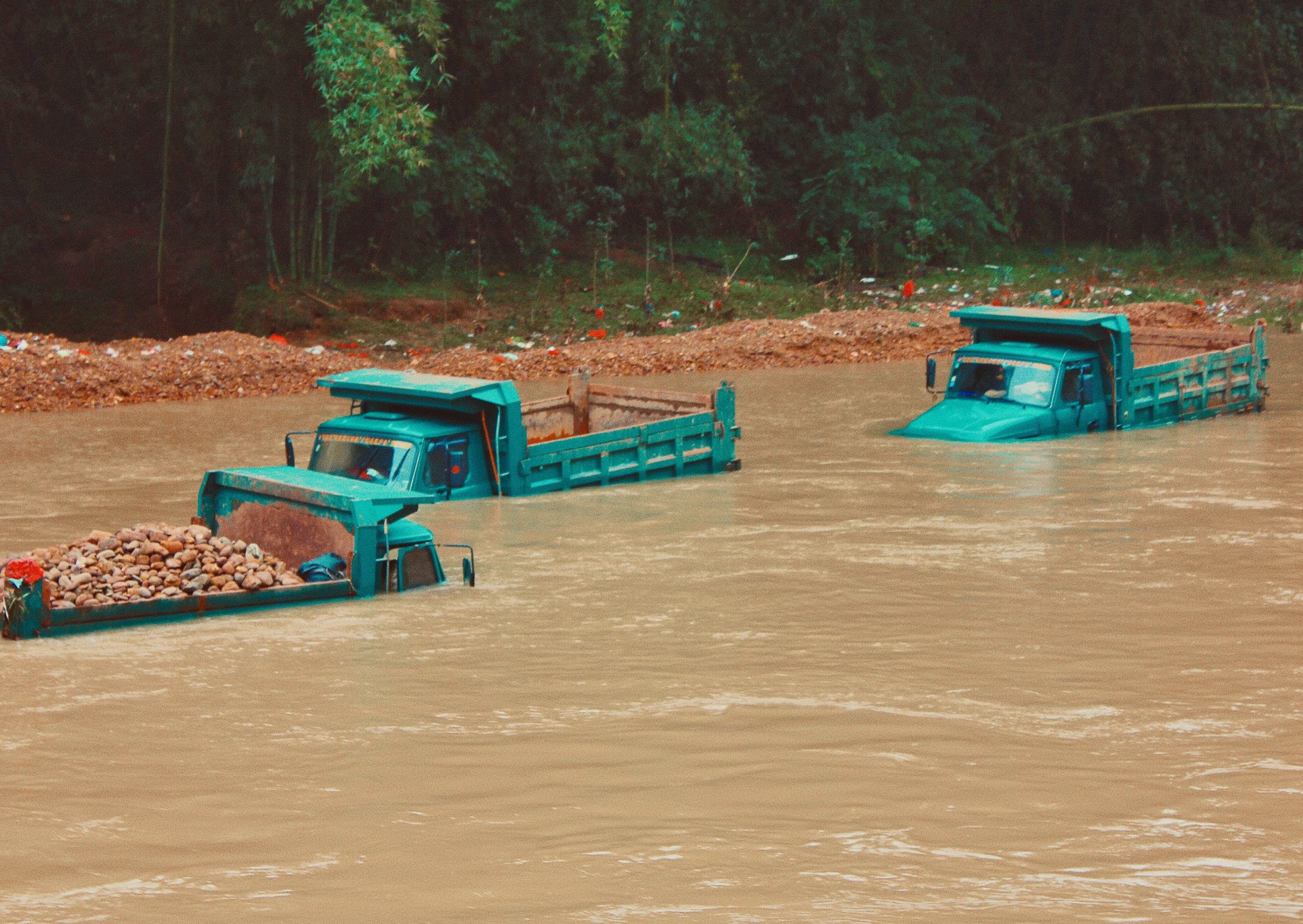 Brown floodwaters swallow three blue trucks by river stop banks and forest.
