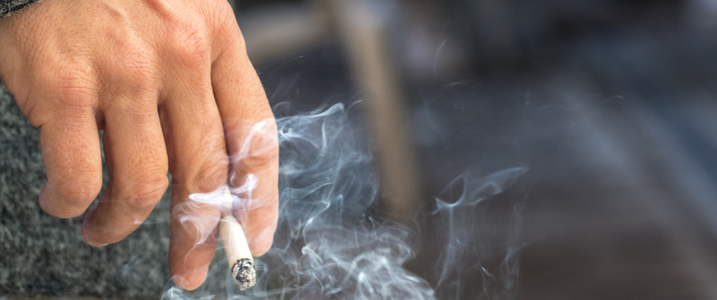 Light-skinned person's hand is shown holding a cigarette with smoke in the air