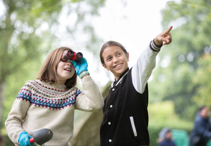 two girls, one pointing and the other looking through binoculars and holding a trowel
