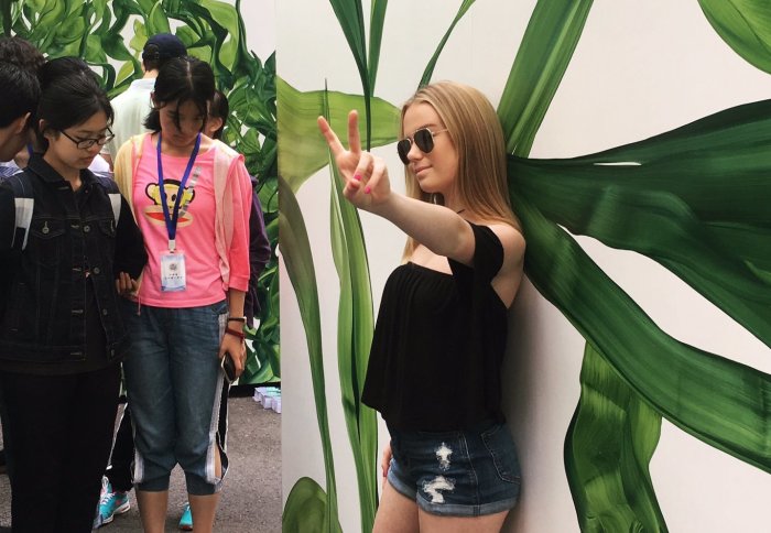 Young lady with round sunglasses poses making a peace sign with her fingers, in front of a walled painted with green foliage