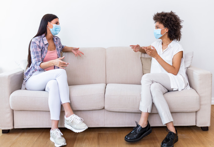 Two female best friends sitting in social distance wearing face mask and talking on the sofa, preventing covid 19 coronavirus pandemic infection spread.