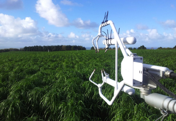 Photo shows a white mechanical arm with finger-like protrusions  held out in front of a field of grasses, it's fine weather with blue sky and fluffy clouds