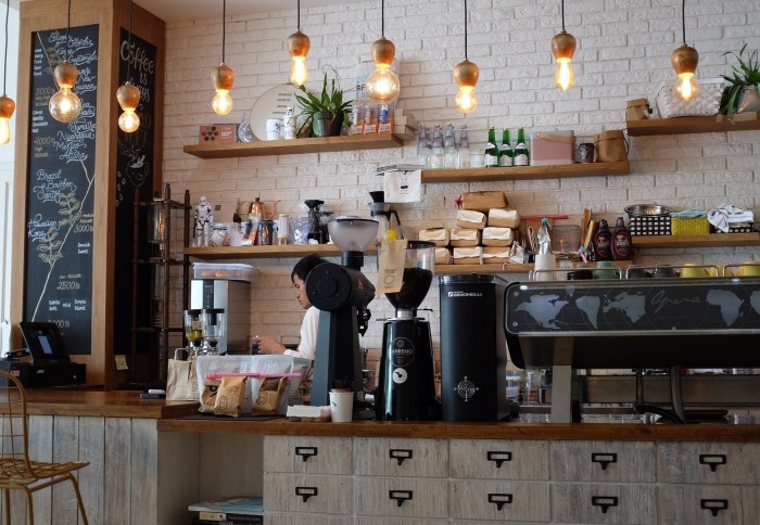 Image of a coffee shop with lightbulbs hanging from the ceiling, coffee making equipment on a wooden countertop and a man in the background making coffee.