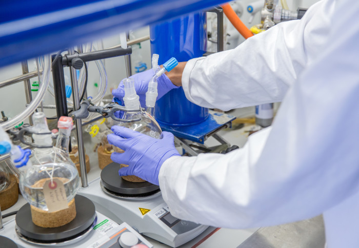 In a lab a person wearing a white labcoat and blue gloves holds a container of liquid on a set of scales