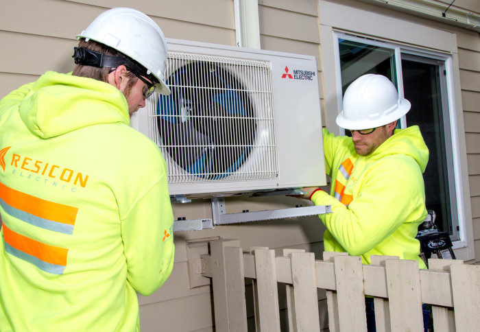 Photo shows two men lifting a heat pump onto brackets on the side of a house. The heat pump is a box about 1m x 0.7m x 0.5m with a grille on the front and a fan inside. The men wear high-viz jackets