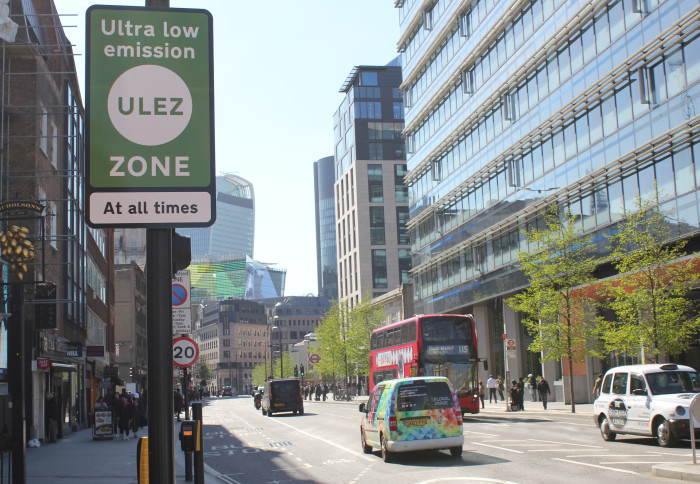 Photo of ULEZ road sign in foreground; red London bus and taxi on road in background