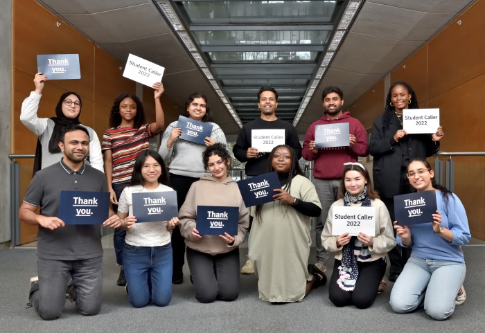 A group of students that are seated and standing smile at the camera and hold signs saying 'thank you' and 'student caller 2022'.