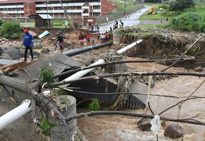 People cross a torrent of muddy flood water on a bank of debris and broken branches