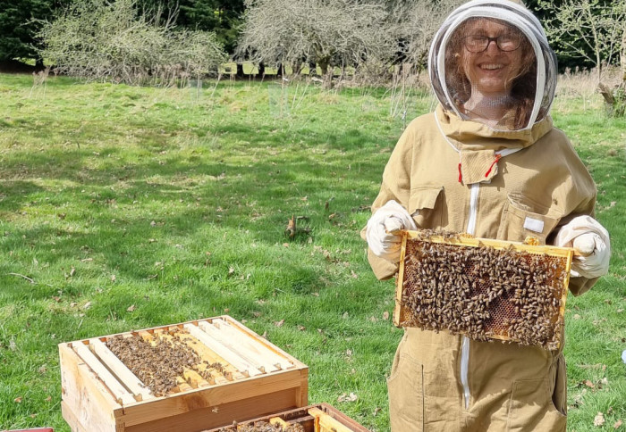 Woman in a beekeeper suit holds up a panel of bees in front of a hive