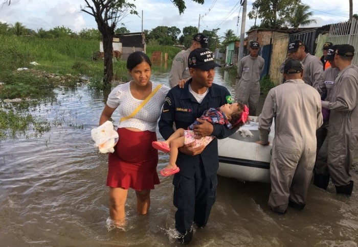 Pregnant woman and her partner carrying a child wade through water surrounded by rescue staff.