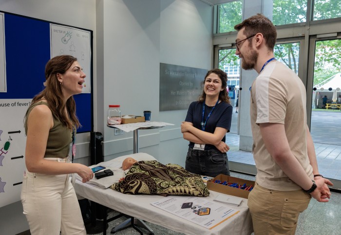 Three researchers stand in conversation at the Great Exhibition Road Festival around an exhibit showing how bed-side tests can be used to diagnose malaria