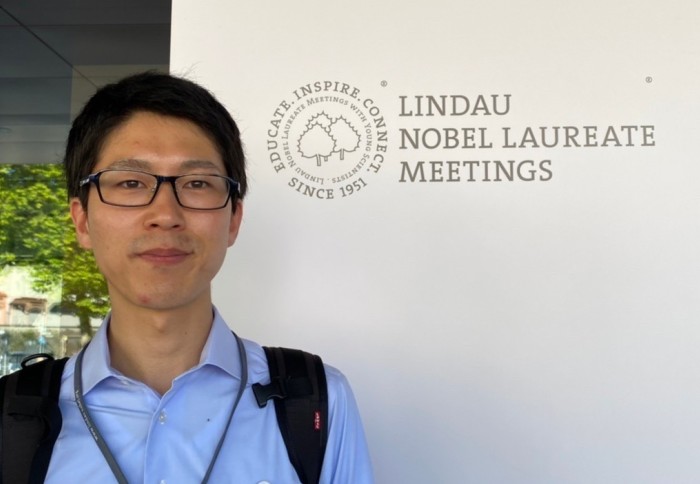 Dr Koichi Sasaki in front of a plaque reading 'Lindau Nobel Laureate Meetings'