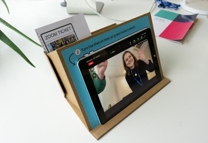 Women on computer tablet waving, with the tablet placed on a cardboard stand.