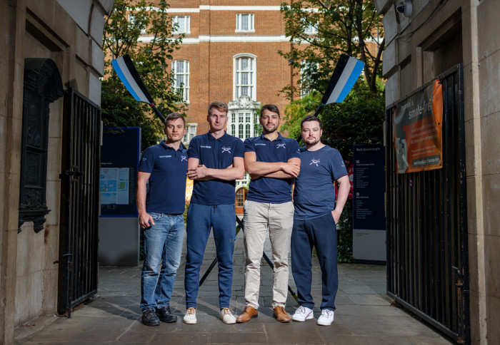 Four men in blue tops stand in front of two rowing oars