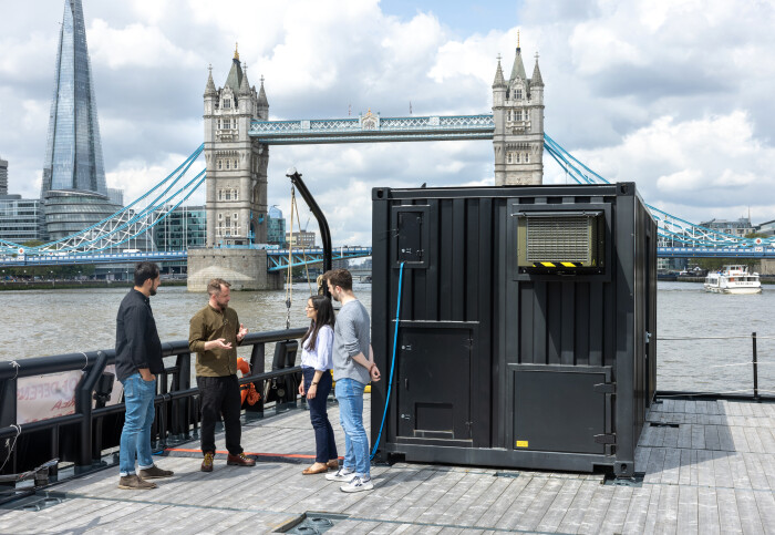 Four people standing outside a black shipping container on the desk of a ship, with Tower Bridge in the background