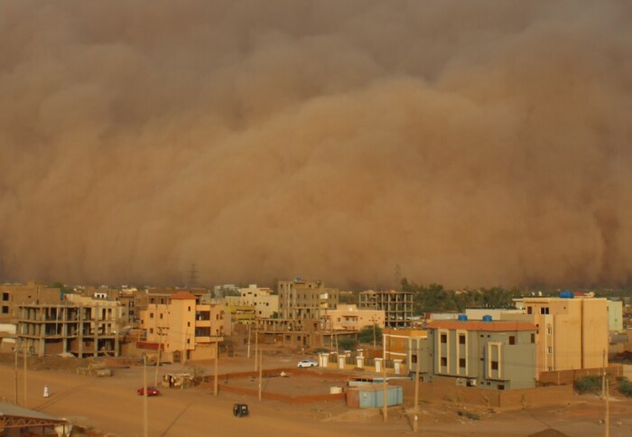Photo of a sandstorm, red desert dust swirls around a lo-rise city.
