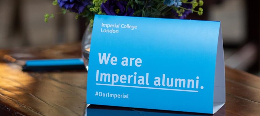 An Imperial branded tent card reading 'We are Imperial alumni #OurImperial' and showing the Imperial logo. The tent card is placed on a table with a backdrop of flowers, indicating the photo was taken at an event