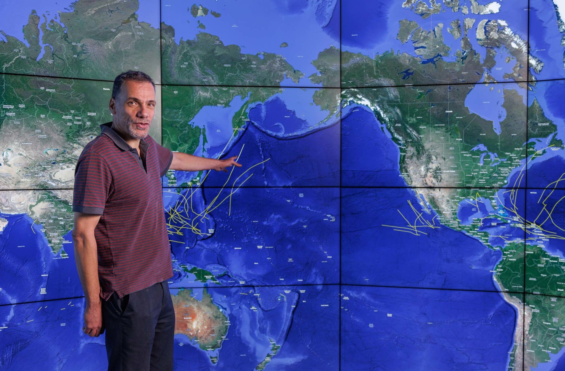 A man pointing to a large screen displaying a map of the world with hurricane tracks drawn on it