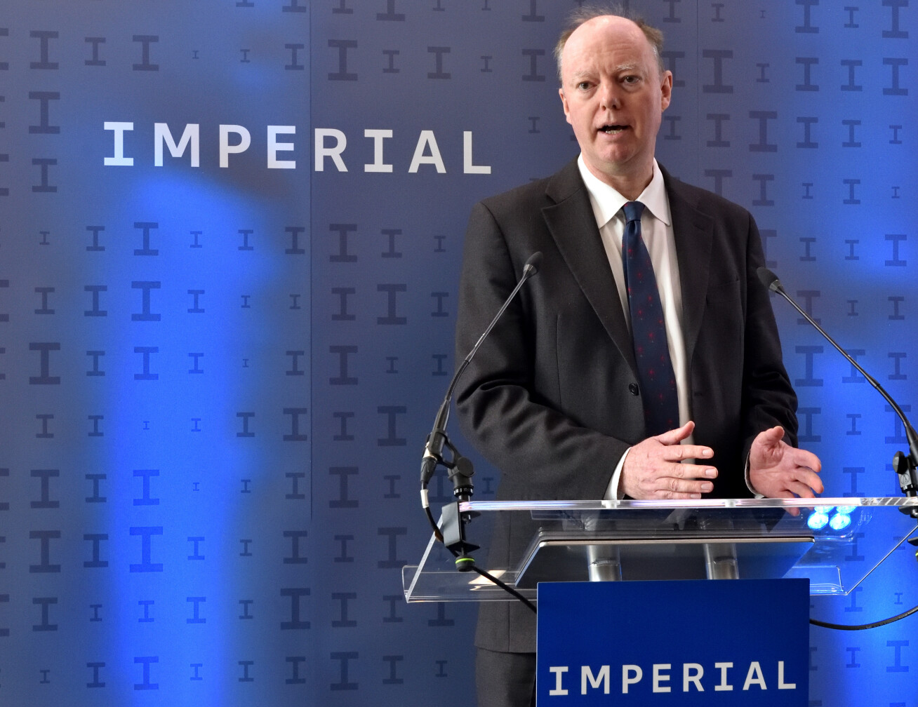Photo: Chris Whitty, a white man in his 50s, speaks from a podium in front of a backdrop saying 'Imperial' in the university's new branding
