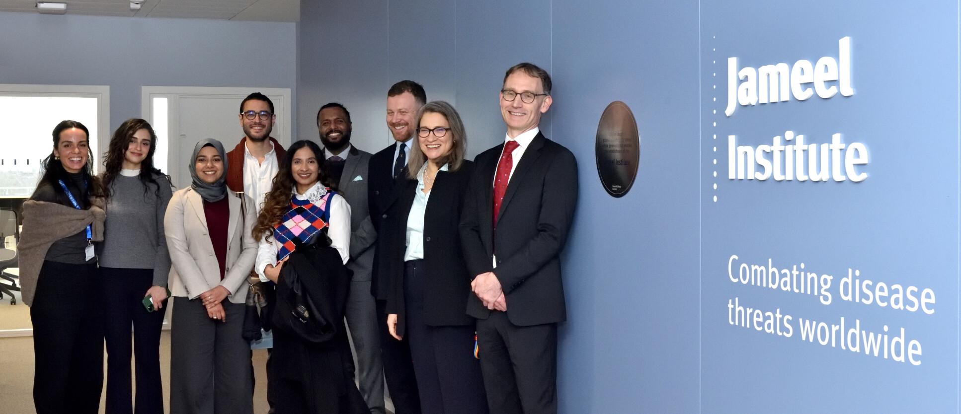 9 smartly dressed people stand proudly in their new office with big smiles on their faces. A sign on the wall reads Jameel Institute / Combating disease threats worldwide.