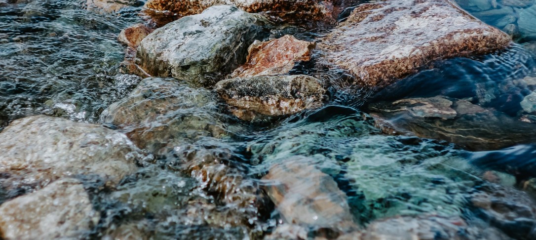 Photo of water running over rocks in the shallow area of a river. By pure-julia on Unsplash