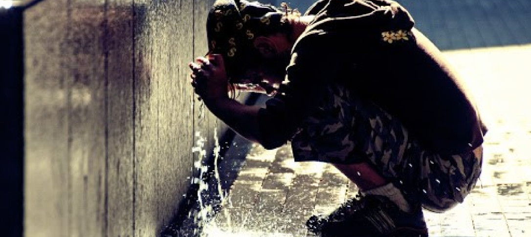 Photo shows a person crouching down by a tap with their head under the water